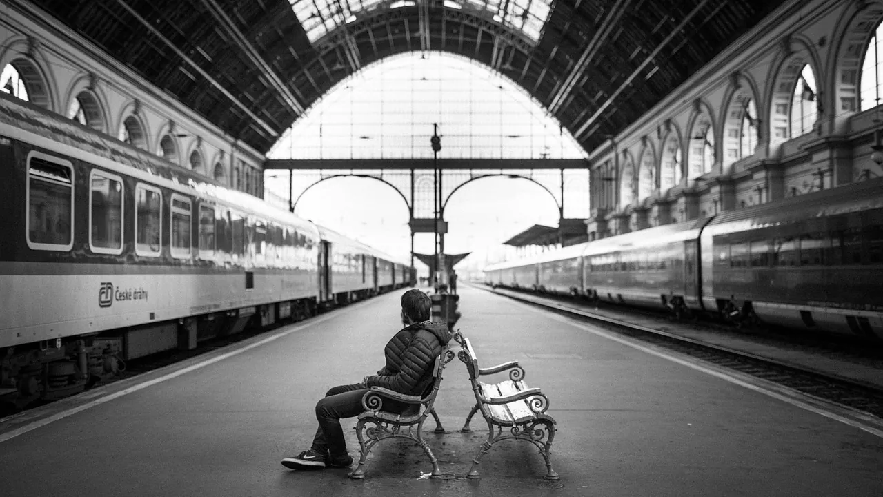 Homme assis seul sur un banc de quai, entre deux trains, sous la verrière d’une gare